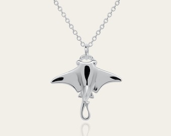 Könnte beinhalten: Halskette mit silbernem Anhänger in Form eines Mantarochens. Der Anhänger hat eine polierte Oberfläche mit schwarzen Akzenten auf den Flügeln. Die Kette ist filigran und aus Gliedern gefertigt. Der Hintergrund ist hell.