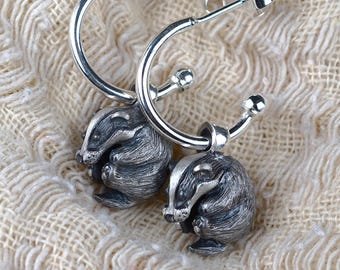 May include: A pair of silver hoop earrings with detailed badger charms. The badger charms are dark gray with white stripes on their faces. The earrings are displayed on a cream-colored, textured fabric.