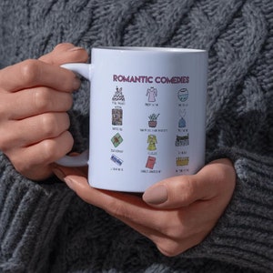 May include: White ceramic mug featuring the text "Romantic Comedies" in purple, with various small illustrations. The mug is held by a person wearing a gray sweater. The illustrations include a cake, a dress, and a fishbowl.