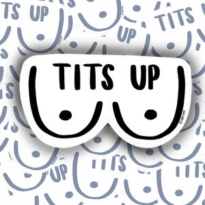 May include: A white sticker with the text "TITS UP" in black, designed to resemble a pair of breasts. The sticker is set against a background of repeating, stylized breast illustrations in light blue, creating a playful and bold design.