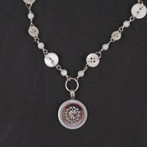 May include: A silver necklace featuring a pendant with a central clear crystal surrounded by a pink and silver border. The necklace chain incorporates white buttons and small, light blue beads.