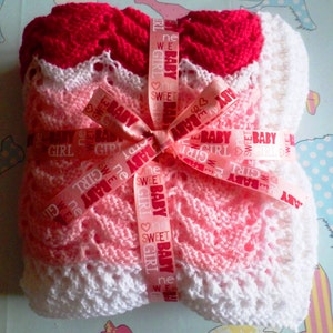 May include: A pink and white knitted baby blanket with a red knitted border. The blanket is tied with a pink ribbon that says "Sweet Baby Girl" repeated on it.