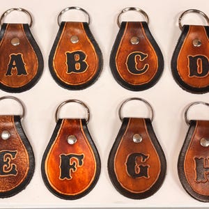May include: Eight leather keychains, each with a different letter from A to H. The keychains are teardrop-shaped with a silver metal keyring. The letters are in a bold, black font, and the leather has a brown and tan color.