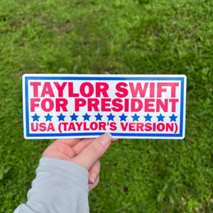 May include: A rectangular bumper sticker with a white background and a blue border. The text reads TAYLOR SWIFT FOR PRESIDENT in red, followed by a row of blue stars and the text USA (TAYLOR'S VERSION) in red at the bottom of the sticker.
