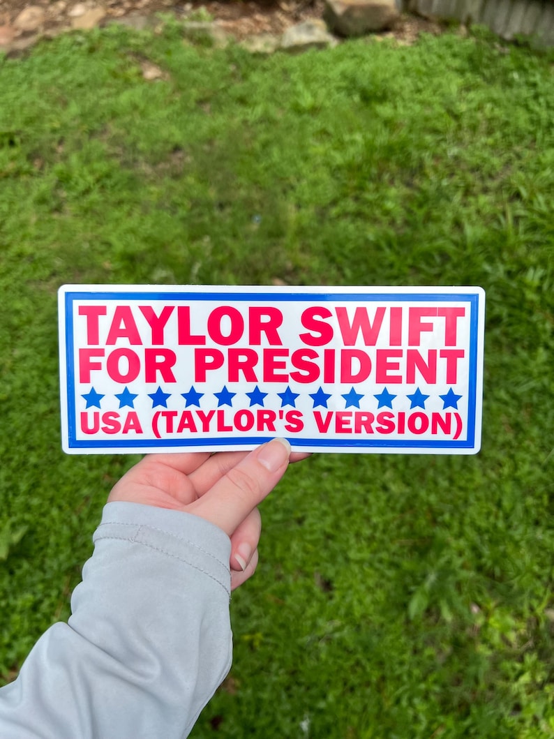 May include: A rectangular bumper sticker with a white background and a blue border. The text reads TAYLOR SWIFT FOR PRESIDENT in red, followed by a row of blue stars and the text USA (TAYLOR'S VERSION) in red at the bottom of the sticker.