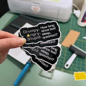 May include: A stack of black and white stickers with the text "Grumpy. Angry. Stupid. How long since last sleep, question?" are held in front of a Cricut machine. The stickers have a white border and a cloud-like shape.