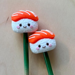 May include: Two decorative sticks topped with miniature sushi figures. Each sushi piece features a white rice base with a smiling face, pink cheeks, and an orange salmon slice on top. The sticks are green at the top and fade to a natural wood color.
