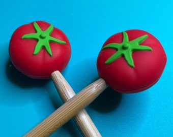 May include: Two wooden knitting needles with red tomato-shaped ends. The needles are crossed and the tomato ends are facing the camera.
