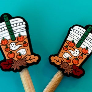 May include: Two wooden knitting needles with orange and brown pumpkin spice latte themed toppers. The toppers feature a white cup with a green straw, orange pumpkins, and white ghosts.