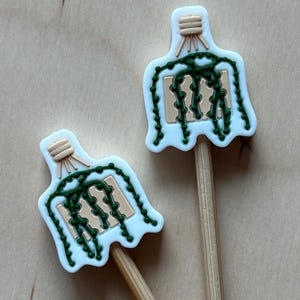 May include: Two plant-themed decorative picks. Each pick features a white, scalloped-edged design with a green plant illustration and a beige wooden stick. The picks are ideal for adding a touch of nature to floral arrangements or crafts.