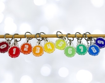 May include: Nine colorful plastic stitch markers with numbers 0 through 8, attached to a wooden dowel rod. The markers are arranged in a rainbow pattern, with red, orange, yellow, green, blue, indigo, and violet.