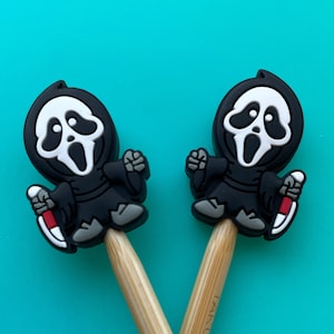 May include: Two wooden knitting needles with black and white Ghostface character toppers. The needles are crossed and the Ghostface characters are facing each other.