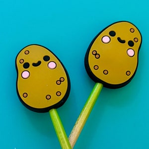 May include: Two wooden knitting needles with yellow and green tips. The tips are shaped like potatoes with brown eyes and pink cheeks. The needles are crossed and the text "Jimmy Beans Wool" is visible on one of the needles.