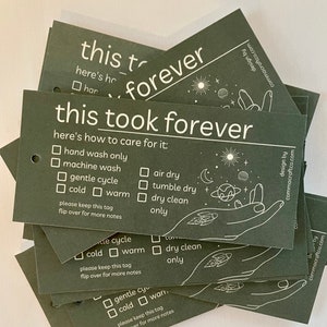 May include: A stack of dark green care tags with white text. The tags have the text "this took forever" and instructions on how to care for the item. The instructions include options for hand wash, machine wash, gentle cycle, cold, warm, air dry, tumble dry, dry clean, and only. The tags also have a small illustration of a hand with a rocket ship.