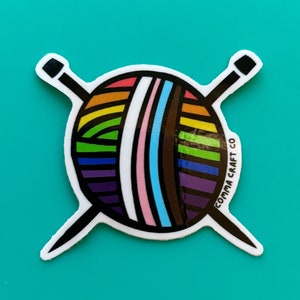 May include: A sticker featuring a colorful yarn ball with rainbow stripes and two black knitting needles crossed behind it. The text "COMMA CRAFT CO" is printed on the right side of the sticker. The sticker is on a teal background.
