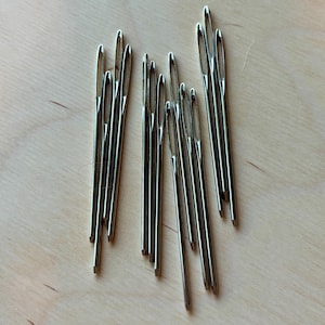 May include: A group of silver sewing needles on a light wood surface.
