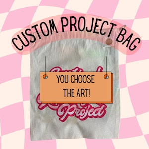 May include: A white cotton tote bag with pink trim. The bag has a pink and white logo that says "Custom Project Bag" and a smaller orange label that says "You Choose The Art!"