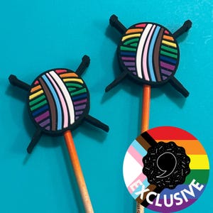 May include: Two rainbow yarn ball pencil toppers with black knitting needles. The yarn balls are made of silicone and have a rainbow color scheme with a white, brown, and pink stripe. The pencil toppers are on a blue background with a sticker that says "EXCLUSIVE" in black text on a rainbow background.