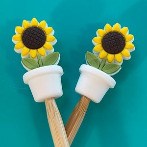 May include: Two wooden knitting needles with sunflower-shaped tops. The tops are white with yellow and brown sunflowers. The needles are crossed.