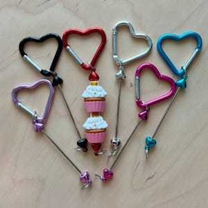 May include: A collection of heart-shaped carabiner clips in various colors, including black, red, silver, blue, pink, and purple. Some clips have decorative cupcake charms. The clips are arranged on a light wooden surface.