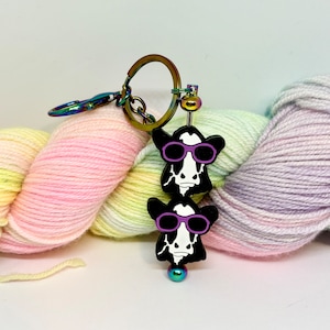May include: A black and white cow keychain with purple sunglasses. The keychain is attached to a rainbow yarn skein.