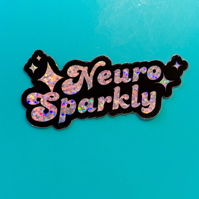 Op de afbeelding: Een zwarte en zilveren holografische sticker met de tekst "Neuro Sparkly" in een retro-lettertype. De sticker heeft een glinsterende afwerking en twee kleine sterren.
