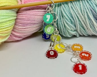 May include: A set of colorful, numbered stitch markers for knitting or crocheting. The markers are attached to a silver chain and feature a rainbow of colors, including red, orange, yellow, green, blue, and purple. The numbers 0 through 7 are visible on the markers.