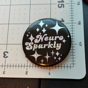 May include: A black button with a white glittery design that says "Neuro Sparkly" with white stars around the text.