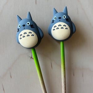 May include: Two gray Totoro-shaped toppers with black eyes, whiskers, and a cream-colored belly, attached to green and tan sticks. The toppers are a cute, whimsical design, perfect for a themed party or as a unique gift.