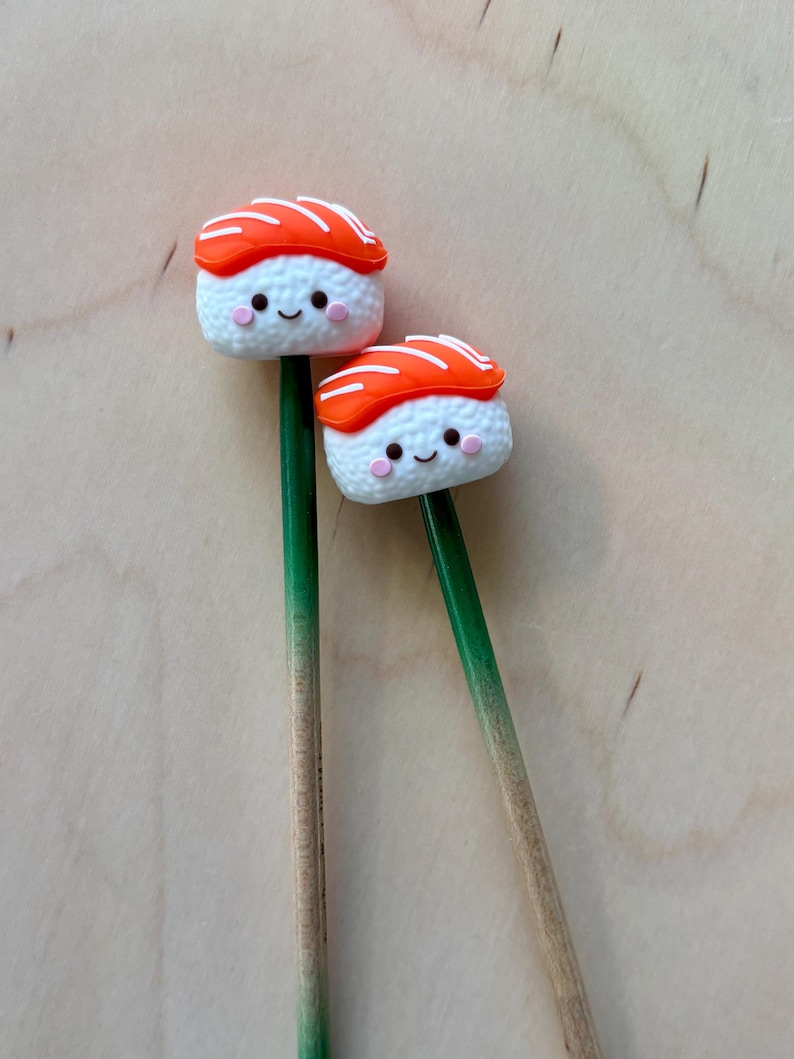 May include: Two decorative sticks topped with miniature sushi figures. Each sushi piece features a white rice base with a smiling face, pink cheeks, and an orange salmon slice on top. The sticks are green at the top and fade to a natural wood color.