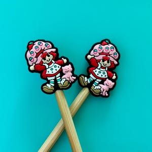 May include: Two wooden knitting needles with strawberry shortcake character toppers. The characters are red, pink, and white with black outlines. The characters are holding a small pink pig.