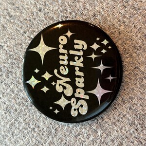 May include: A black button with a white glittery design that says "Neuro sparkly".