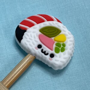 May include: A silicone sushi-shaped charm featuring a white rice texture, a red and white fish topping, and colorful vegetable accents. The charm has a small, smiling face with black eyes and a pink blush, and it is attached to two wooden sticks.