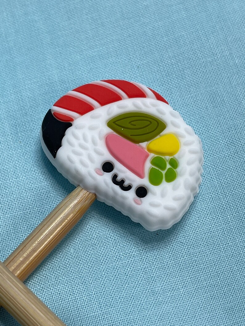 May include: A silicone sushi-shaped charm featuring a white rice texture, a red and white fish topping, and colorful vegetable accents. The charm has a small, smiling face with black eyes and a pink blush, and it is attached to two wooden sticks.