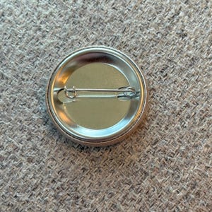 May include: The back of a round button with a silver pin and a silver clasp. The button is made of metal and has a smooth, shiny surface.