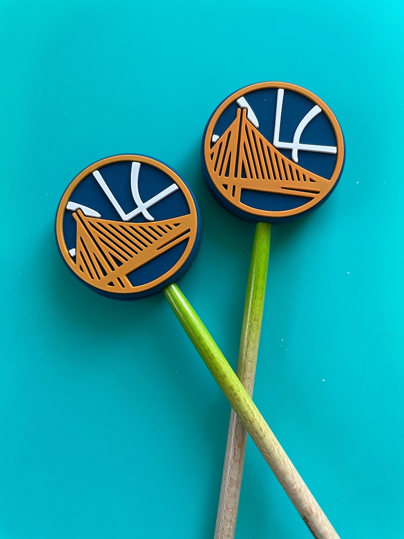 May include: Two wooden pencils with blue and gold round toppers featuring the Golden State Warriors logo. The logo includes a basketball and the Golden Gate Bridge.