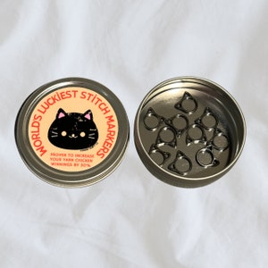 May include: A round metal tin with a black cat illustration and the text "World's Luckiest Stitch Markers". The tin contains a set of black cat-shaped stitch markers.
