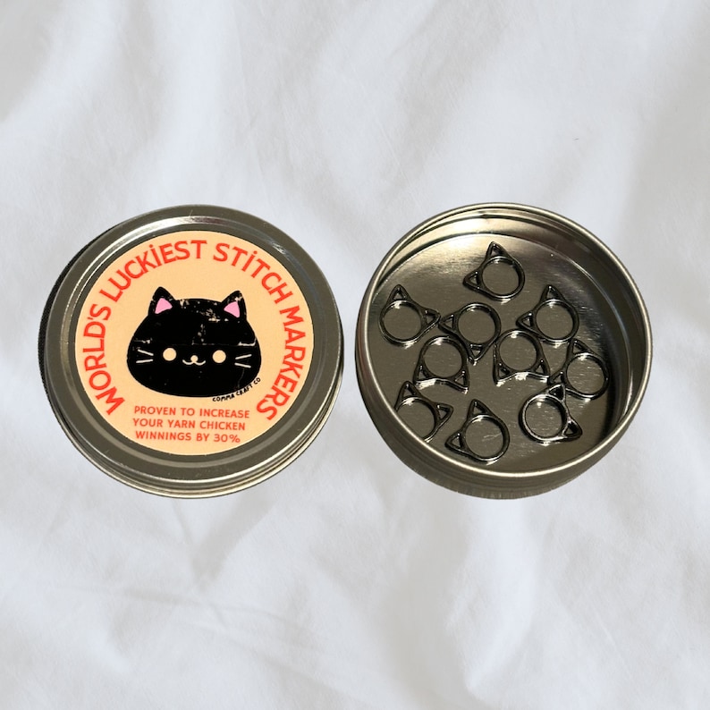 May include: A round metal tin with a black cat illustration and the text "World's Luckiest Stitch Markers". The tin contains a set of black cat-shaped stitch markers.