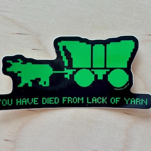 May include: A die-cut sticker featuring a bright green pixelated graphic of an ox pulling a covered wagon against a black background. Below the graphic, the text reads in green pixelated font: YOU HAVE DIED FROM LACK OF YARN.