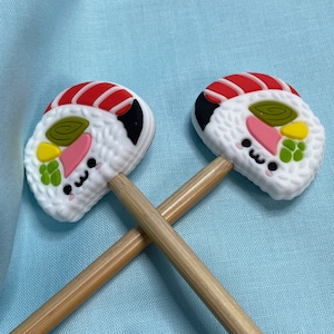 May include: Two wooden pencils topped with silicone sushi roll toppers. Each topper features a white rice base, a red and white striped fish slice, a green swirl, a yellow piece, and a pink slice, all with a small black kawaii face.