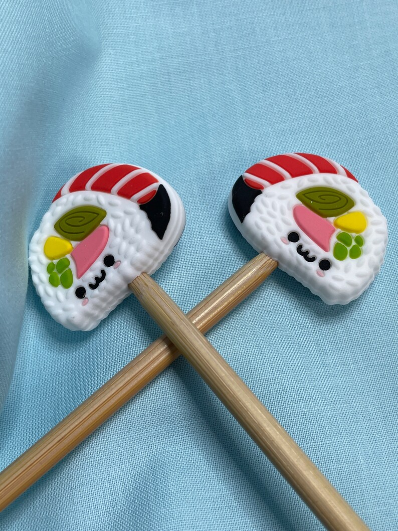 May include: Two wooden pencils topped with silicone sushi roll toppers. Each topper features a white rice base, a red and white striped fish slice, a green swirl, a yellow piece, and a pink slice, all with a small black kawaii face.