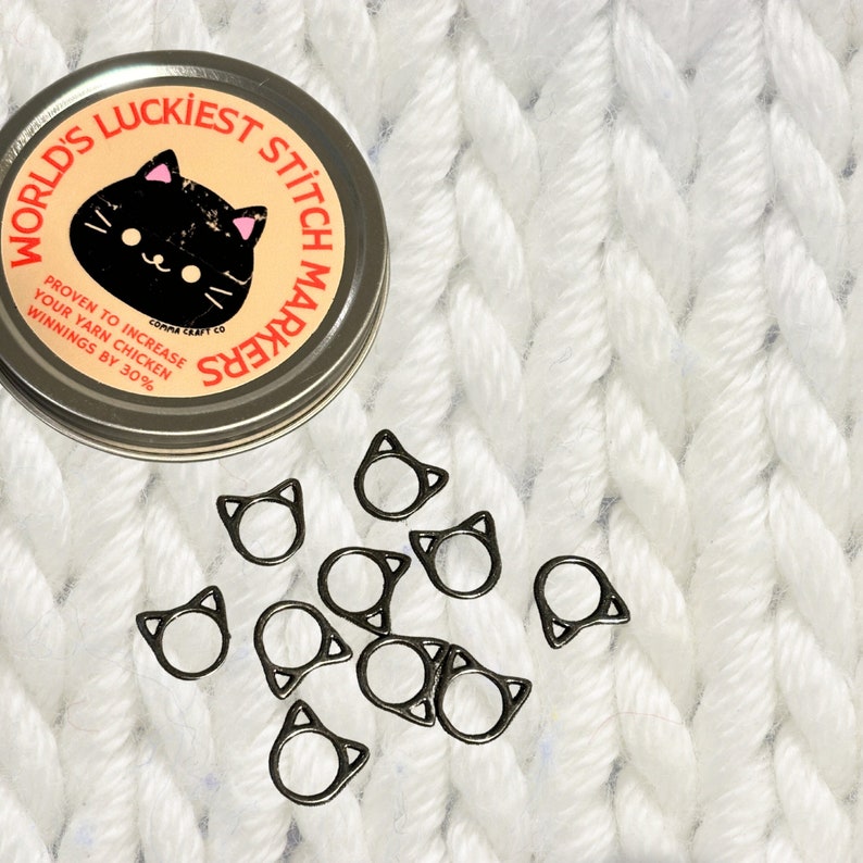 May include: A tin of black cat stitch markers with the text "World's Luckiest Stitch Markers" and "Proven to increase your yarn chicken winnings by 30%". The tin is open and the markers are scattered on a white knitted surface.