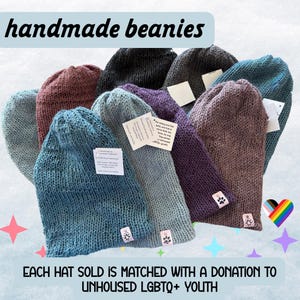 May include: A collection of handmade knit beanies in various colors including blue, purple, maroon, black, and gray. Each hat features a small tag with a paw print or geometric design and a white product information label attached to the fabric.