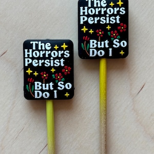 May include: Two black rectangular signs with the text "The Horrors Persist But So Do I" in white. The signs have yellow stars and red flowers. The signs are attached to yellow sticks.