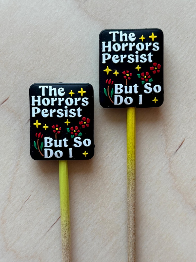 May include: Two black rectangular signs with the text "The Horrors Persist But So Do I" in white. The signs have yellow stars and red flowers. The signs are attached to yellow sticks.
