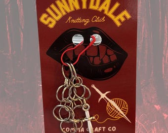 May include: A red card with the text "SUNNYDALE Knitting Club" and a graphic of a mouth with fangs. A silver chain with a small sword charm hangs from the card. The card also features the text "COMMA CRAFT CO" and a website address.