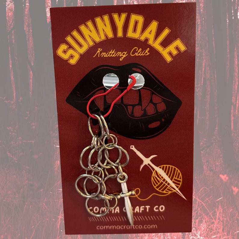 May include: A red card featuring the text "SUNNYDALE Knitting Club" and a mouth graphic with fangs. A silver chain with a small sword charm is attached. The card also displays "COMMA CRAFT CO" and a website address.