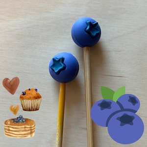 May include: Two blueberry-shaped cake pops on wooden sticks, with a dark blue star-shaped design on top. The image also includes illustrations of a muffin, pancakes, blueberries, and heart shapes, all on a light wood grain background.