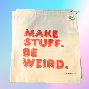 May include: A white canvas tote bag with red text that reads "MAKE STUFF. BE WEIRD." and the words "COMMA CRAFT CO" in smaller text at the bottom. The bag has a green eco-friendly label.