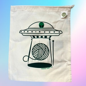 May include: A white canvas tote bag with a green alien spaceship design. The spaceship is hovering over a ball of yarn and knitting needles. The bag has a green "ECO BAGS" label and the text "COMMA CRAFT CO" at the bottom.